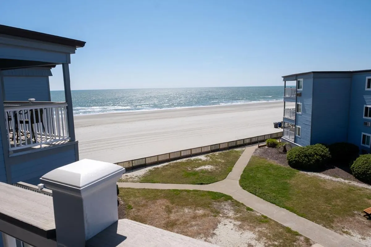 The image showcases a stunning ocean view from the condo, with a sun-drenched sandy beach, gentle waves, and a scenic pathway leading directly from the building to the shoreline. Guests can be seen enjoying sunbathing, walking along the shore, swimming, and families building sandcastles, all set against the serene backdrop of Myrtle Beach's inviting coastline.