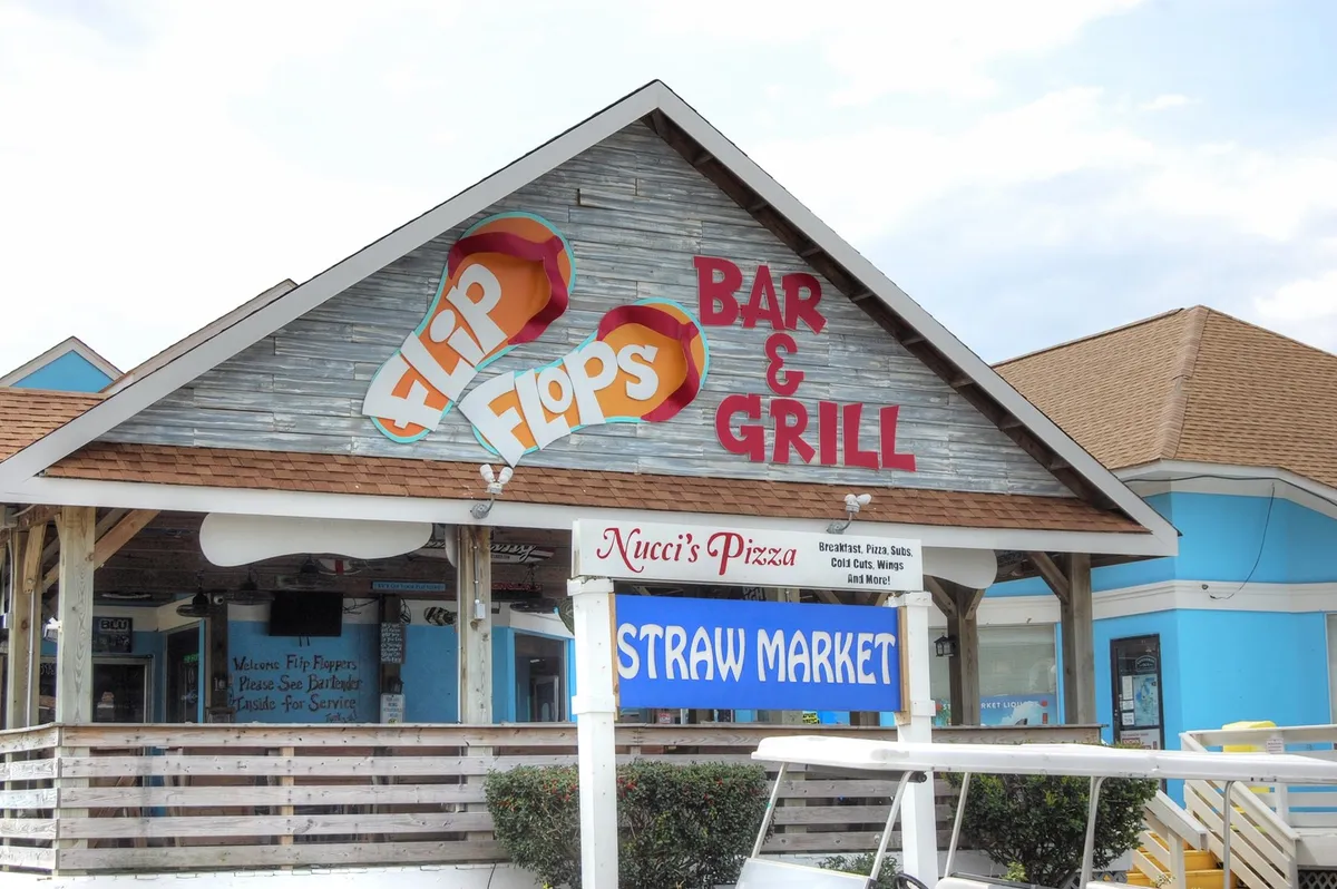 The image captures a lively corner of Shore Drive featuring prominent signage for Flip Flops Bar & Grill, Nucci's Pizza, and the Straw Market. The scene suggests a fun, social atmosphere for dining, music, and mingling with friends—all within walking distance of the condo.