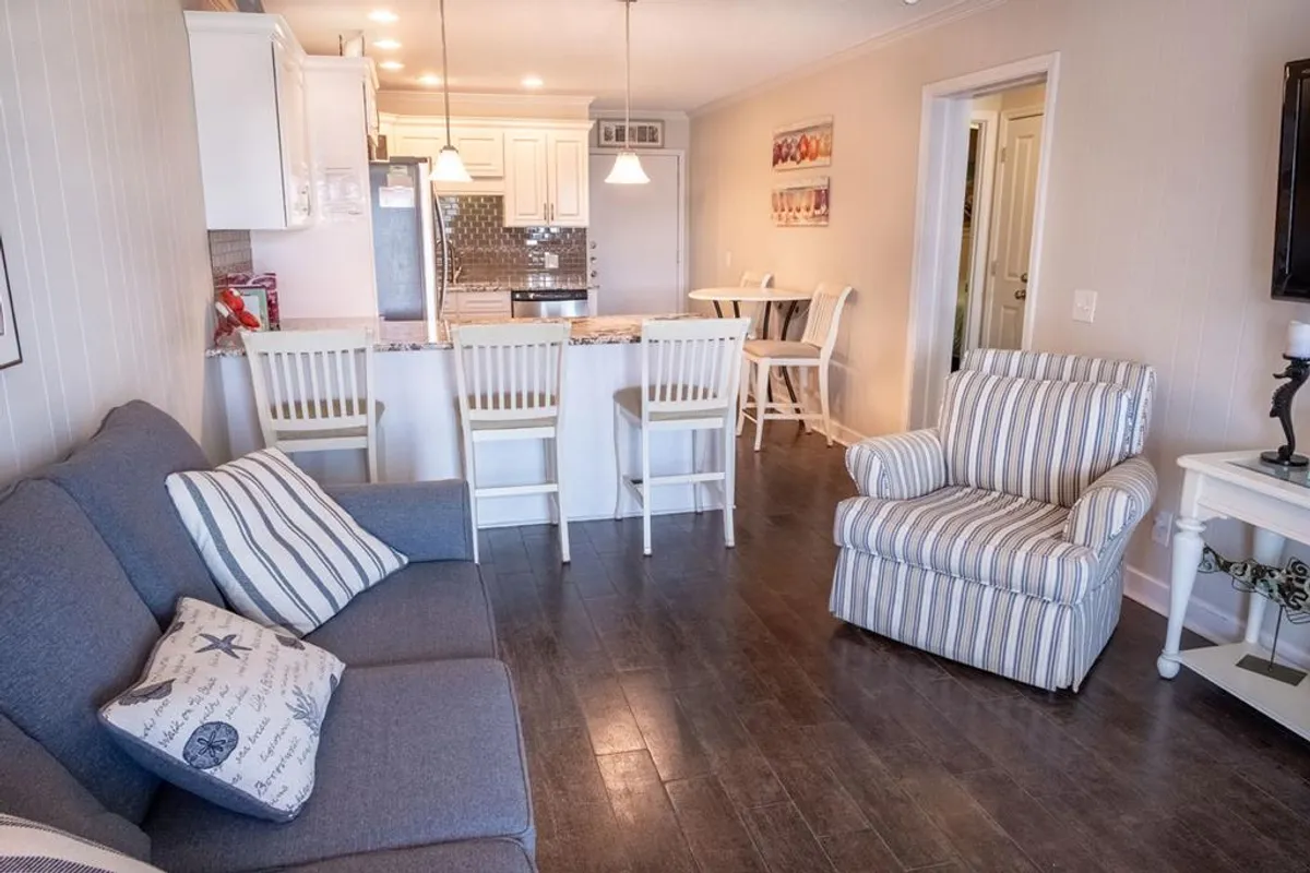 This image captures a sunlit open-plan living and dining area with a cozy sofa adorned with decorative pillows, a striped armchair, a kitchen island with bar stools, and a dining table set for gatherings. Art on the wall and natural light streaming in create an inviting and togetherness-focused environment.
