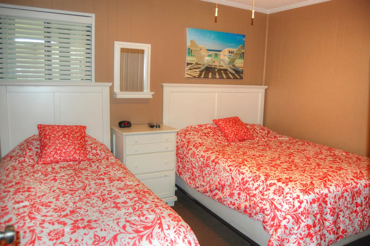 A cheerful bedroom featuring two beds with floral red and white bedding, a white dresser, a mirror, and a painting of a beach scene. The room is bright and welcoming, perfect for accommodating families or groups.