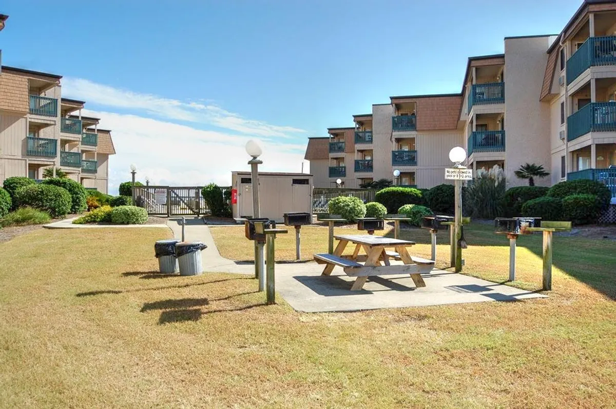 A vibrant outdoor communal area featuring picnic tables and grill stations set among landscaped greenery with the condo’s balconies visible in the background. The area is designed for outdoor meals, barbecues, and socializing in a cheerful, natural setting.