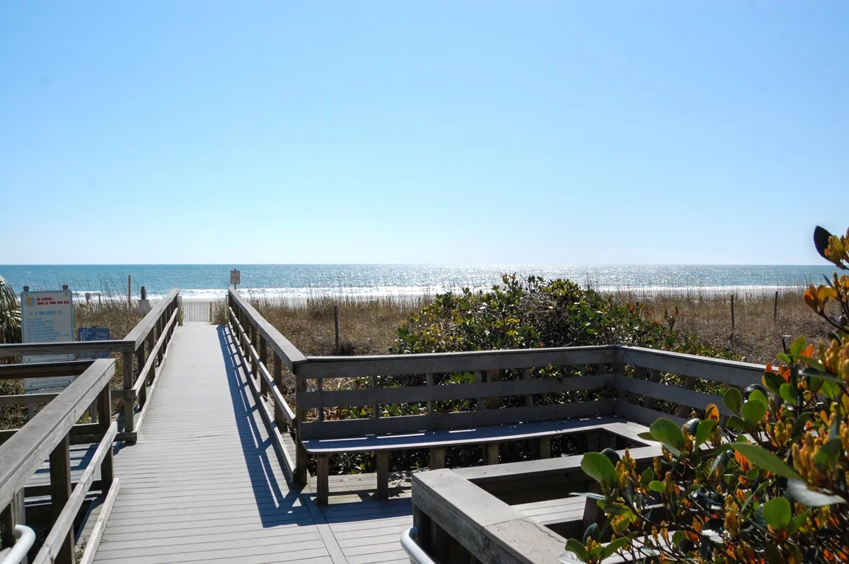 This image captures a sunlit wooden boardwalk leading to the beach, bordered by natural dune vegetation and an expansive view of the ocean beyond. The scene is tranquil, inviting, and emphasizes instant access to the sand and sea, hinting at the start of a fun-filled family day by the water.