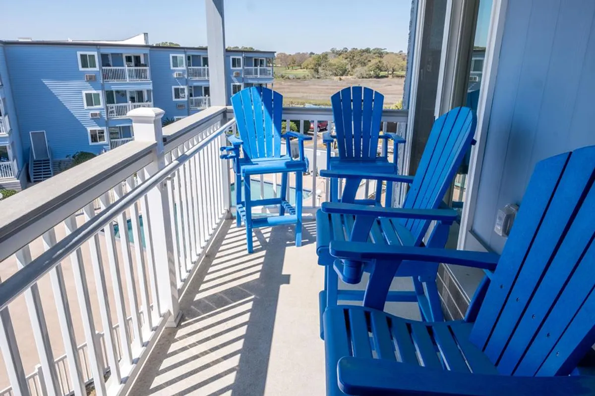 This image captures a serene scene from the private balcony, featuring blue Adirondack chairs arranged to take in the view through white railings. The distant ocean and soft natural light suggest an ideal spot for morning coffee or unwinding at sunset.