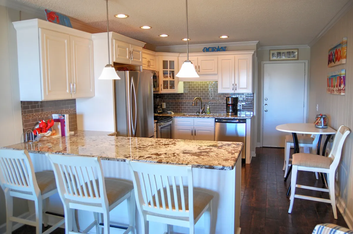 A friendly, open kitchen scene with a large granite countertop, bar stools for casual seating, stainless steel appliances, a central kitchen island, and decorative wall art. The setup encourages socializing and casual meals, ideal for group breakfasts or evening gatherings.