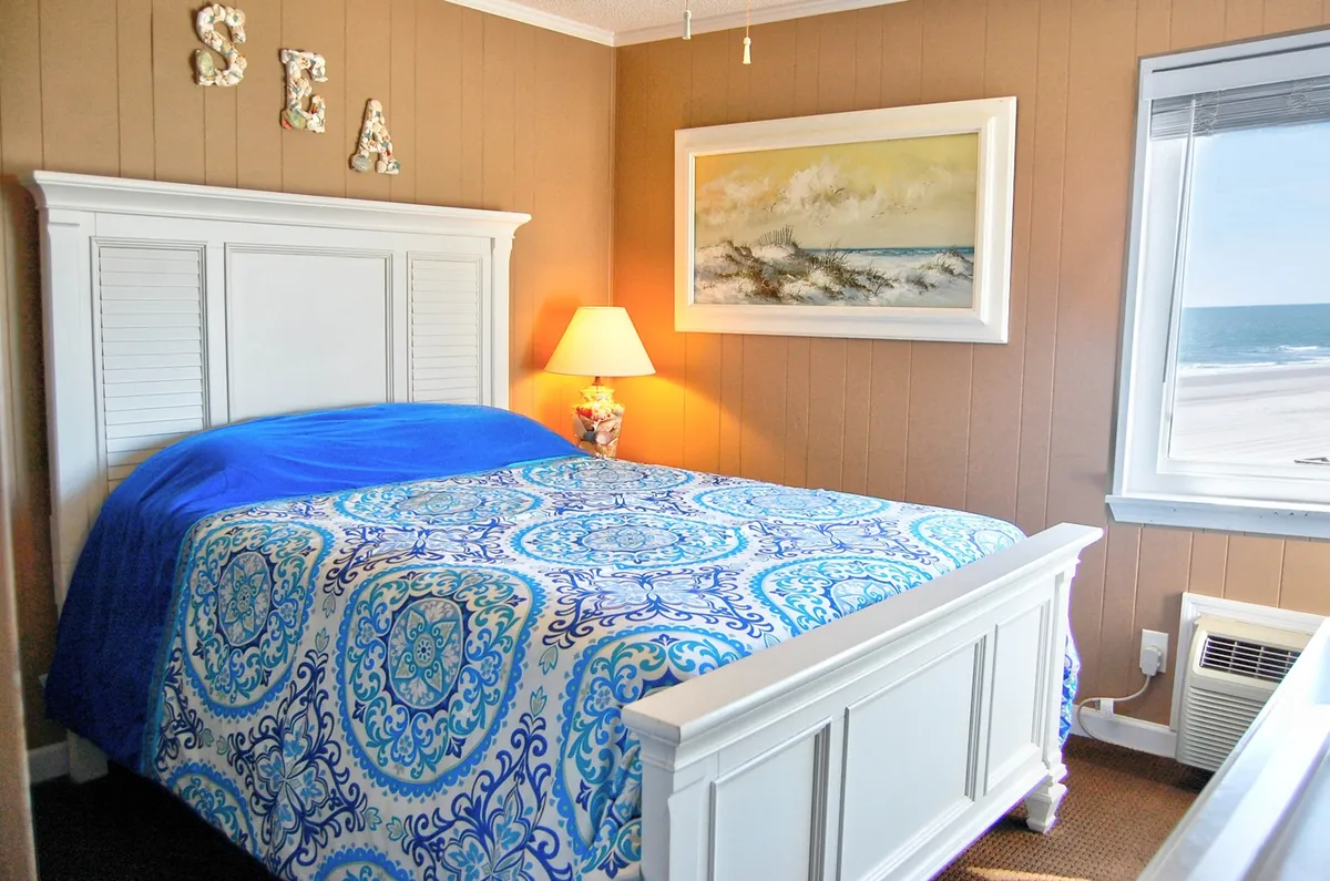 A serene bedroom with a Queen-sized bed adorned in blue and white patterned bedding. The room is softly lit by a lamp, and there is framed artwork of a beach scene on the wall. Natural light filters in through a window, suggesting a peaceful, restful ambiance.