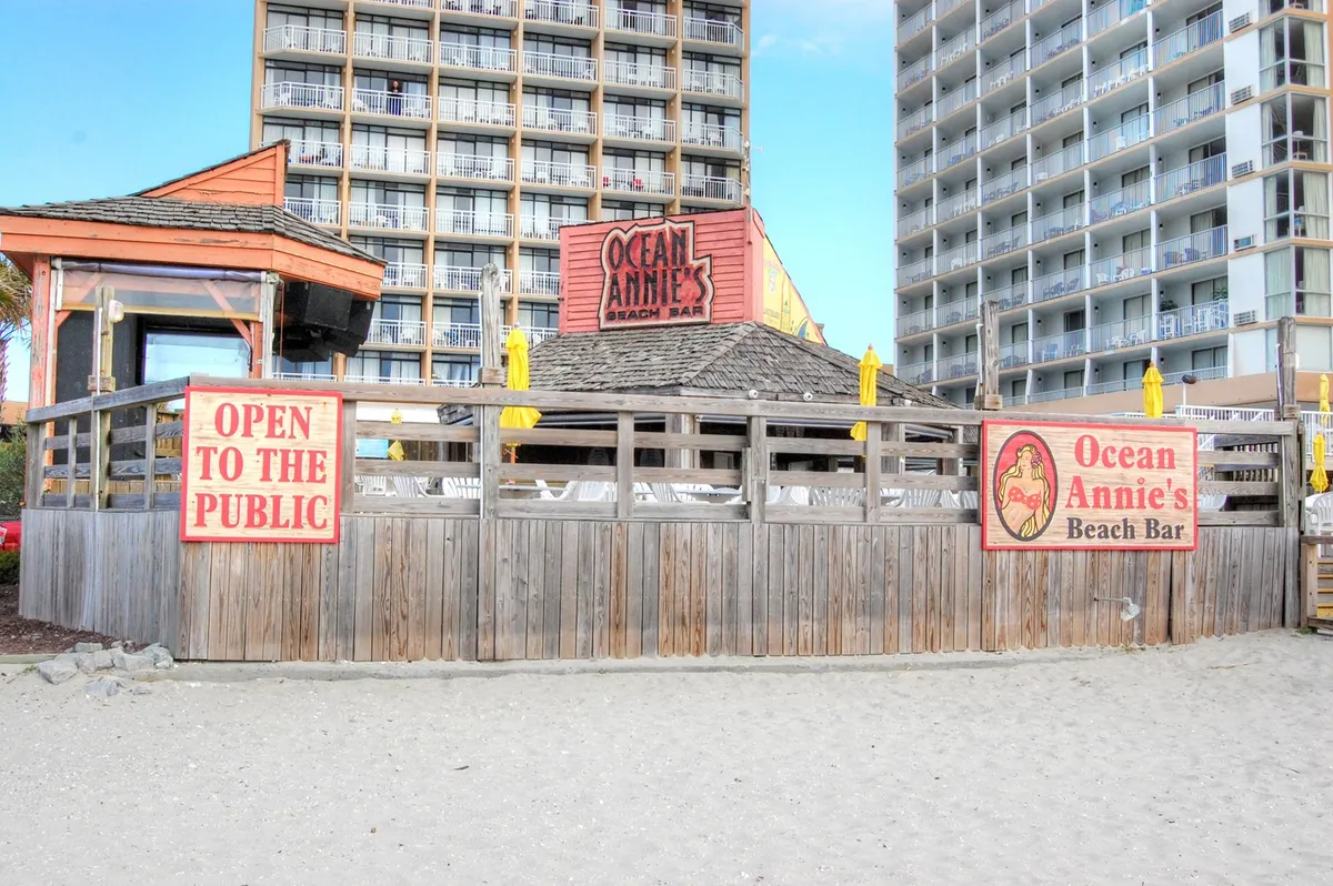 The lively exterior of Ocean Annie's Beach Club, featuring its signature sign, open-to-the-public banner, wooden railing, and cheerful yellow umbrellas. The setting is festive with a hint of beachside relaxation and social activity.