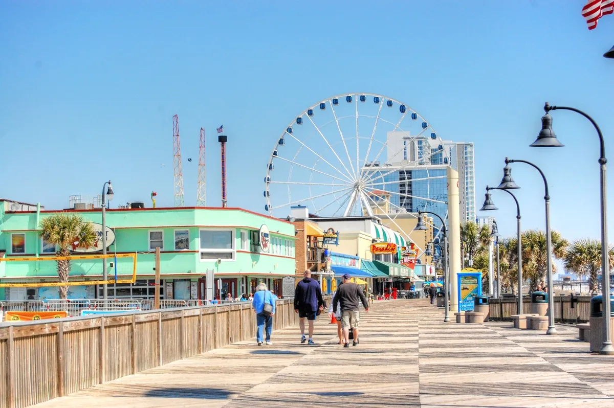 This exterior image features the iconic Myrtle Beach Ferris wheel, palm trees, colorful buildings, and a lively boardwalk. People are seen strolling and enjoying the vibrant atmosphere, representing the area's fun attractions and entertainment options.