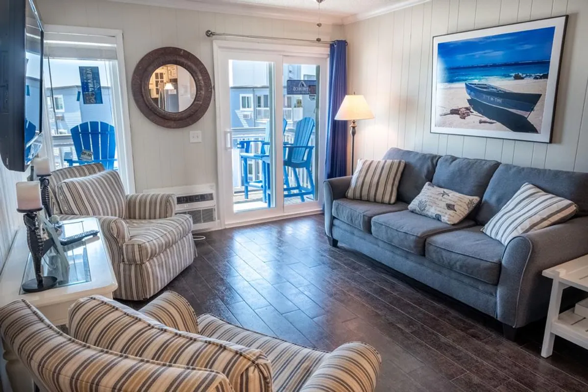 A welcoming living room with a plush grey sofa oriented towards a large wall art featuring a beach scene, with visible blue chairs on the adjacent balcony. The scene hints at easy access to outdoor space and the presence of large windows letting in ample coastal light.