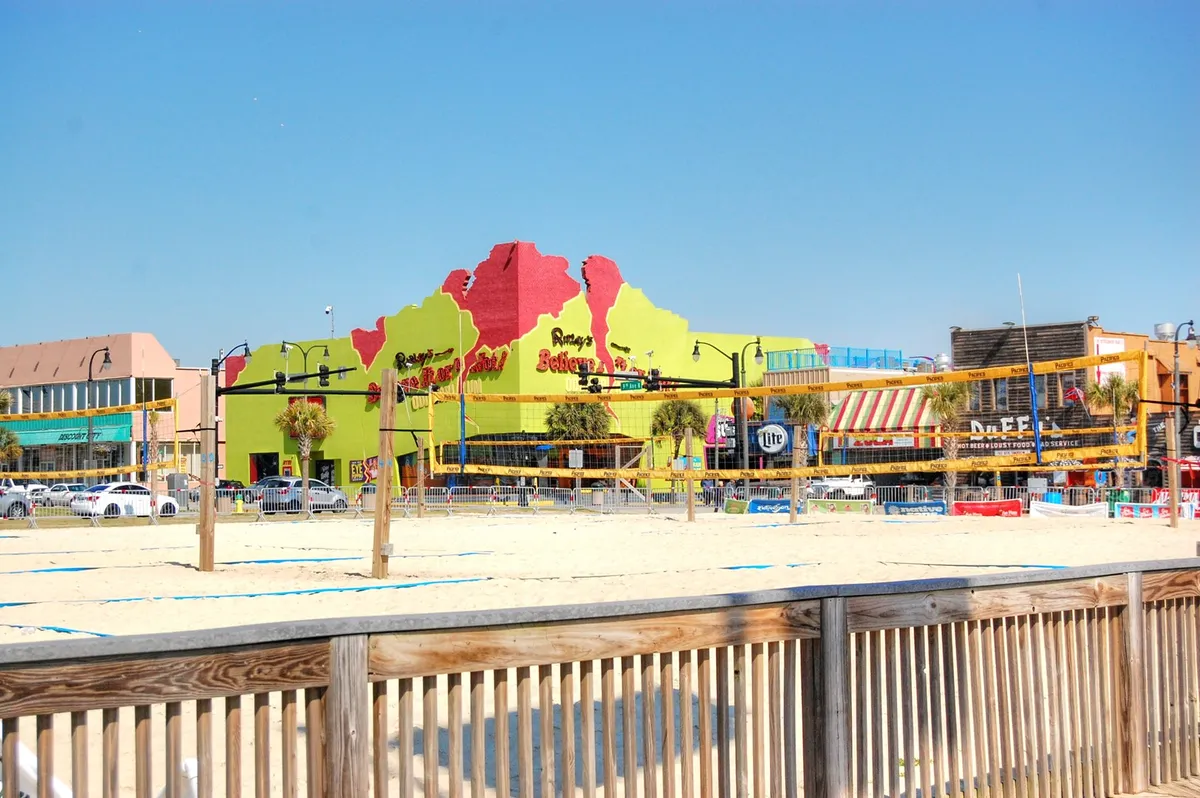 A colorful exterior view of a lively area featuring beach volleyball courts, vibrant building facades, palm trees, and a variety of shops and restaurants. The atmosphere is energetic and sociable, suggesting a place buzzing with local events and family-friendly amenities.