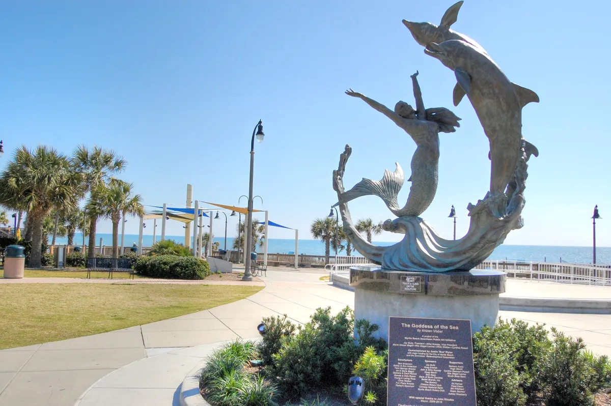 A scenic park with the iconic 'Goddess of the Sea' sculpture, palm trees, ocean views, and park benches along a beachside promenade. The area invites leisurely strolls, relaxation, and enjoying public art against a coastal backdrop.