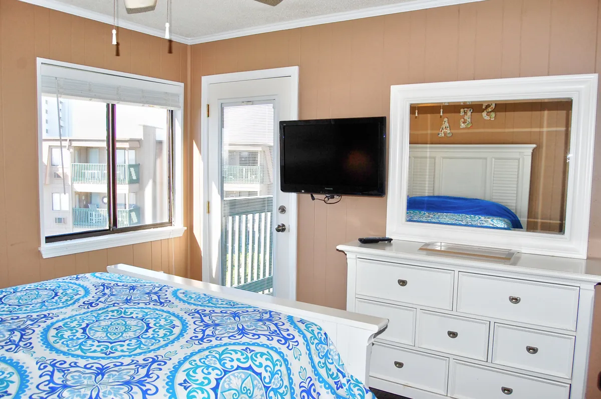 A cozy bedroom interior with a blue patterned comforter and a door leading out to the balcony. A television and a mirror enhance the sense of comfort, while the natural light streaming in emphasizes the welcoming mood.